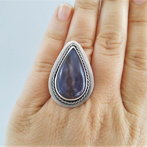 May include: A silver ring featuring a large, teardrop-shaped, polished stone with shades of purple and gray. The stone is set within a silver frame with a twisted rope design around the edge. The ring is displayed on a hand.