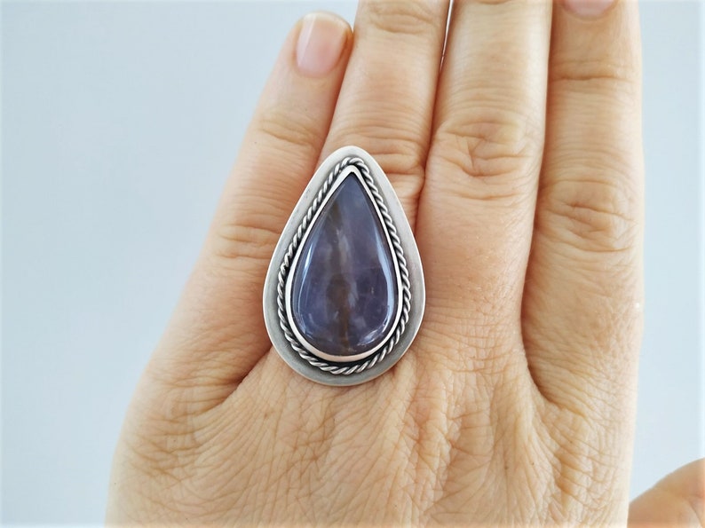 May include: A silver ring featuring a large, teardrop-shaped, polished stone with shades of purple and gray. The stone is set within a silver frame with a twisted rope design around the edge. The ring is displayed on a hand.