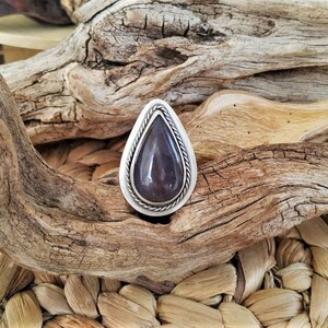 May include: A teardrop-shaped ring with a dark purple stone set in a silver-colored metal. The stone is surrounded by a twisted rope design. The ring is displayed on a textured, natural background of wood and woven fibers.