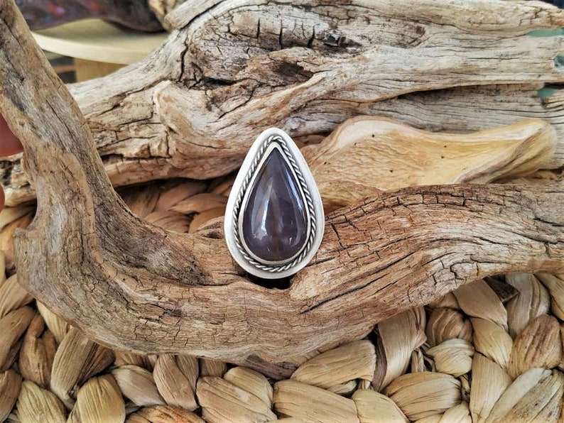 May include: A teardrop-shaped ring with a dark purple stone set in a silver-colored metal. The stone is surrounded by a twisted rope design. The ring is displayed on a textured, natural background of wood and woven fibers.