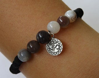 May include: A bracelet with grey, brown, and white agate beads. A silver charm with an Om symbol is attached. The bracelet is strung on a black elastic cord.