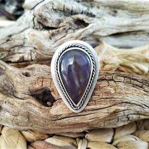 May include: A silver-toned ring with a teardrop-shaped, polished purple stone. The stone is set in a bezel with a twisted rope design around the edge. The ring is displayed on a piece of driftwood.