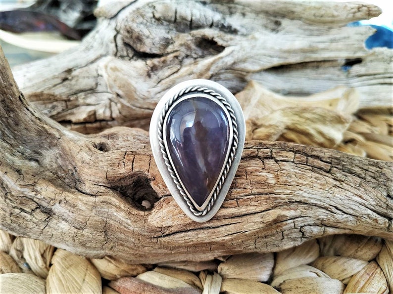 May include: A silver-toned ring with a teardrop-shaped, polished purple stone. The stone is set in a bezel with a twisted rope design around the edge. The ring is displayed on a piece of driftwood.