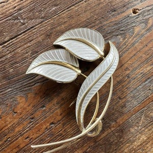 Peut inclure: Broche vintage dorée représentant trois feuilles stylisées avec de l'émail blanc et des accents dorés. Les feuilles présentent des nervures détaillées. La broche est posée sur une surface en bois rustique.