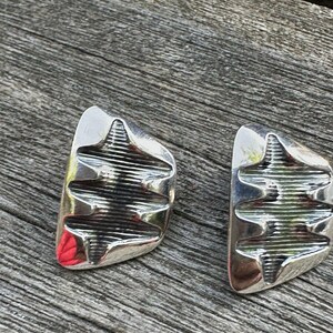 Peut inclure: Une paire de boucles d'oreilles argentées au design géométrique. Chaque boucle d'oreille a une forme rectangulaire incurvée avec une série de rainures horizontales. Les boucles d'oreilles sont présentées sur une surface en bois vieilli.