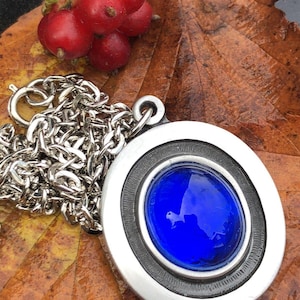 Puede incluir: Un collar con colgante plateado con una gran piedra ovalada de cristal azul oscuro. El colgante tiene un marco circular con un borde negro. La cadena es plateada, con una hoja marrón y bayas rojas en el fondo.