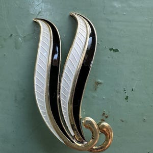 Peut inclure: Broche vintage en forme de feuille. La broche présente un cadre en métal doré avec des détails en émail noir, blanc et or. Le design comprend deux formes incurvées en forme de feuille avec un centre blanc texturé et des bordures noires.