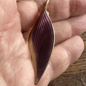 Peut inclure: Broche vintage en forme de feuille. Elle présente un cadre en métal doré et une section centrale en émail violet foncé nervuré. La forme de la feuille est allongée et légèrement incurvée, avec une pointe.
