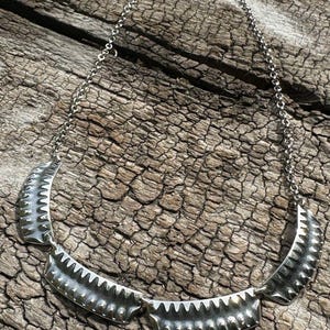 Peut inclure: Un collier argenté au design unique. Le collier est composé d'une chaîne délicate et de plusieurs éléments métalliques incurvés et texturés. Les pièces métalliques ont un bord en dents de scie. Le collier est présenté sur une surface en bois vieilli.