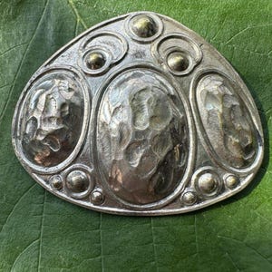 Könnte beinhalten: Silberfarbene Metallbrosche mit abstraktem Design. Die Brosche hat drei strukturierte ovale Formen und mehrere kleinere kreisförmige Akzente. Das Metall hat ein gehämmertes Aussehen, auf einem grünen Blatt.