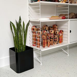 May include: A white wire shelving unit displays a collection of wooden dolls with painted faces and clothing. A black square planter holds a snake plant with green and yellow striped leaves. A yellow smiley face decorates the top shelf.