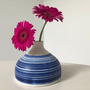 May include: A white ceramic vase with blue stripes, holding two pink gerbera daisies.