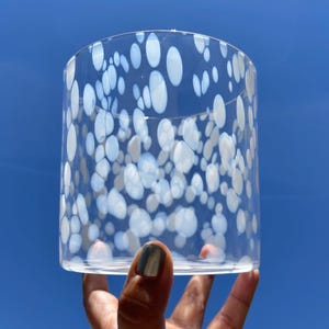 May include: A clear glass cylinder with a speckled pattern of white and light blue ovals. The glass is held against a bright blue sky, showcasing the design. The cylinder is likely a decorative item or a container.