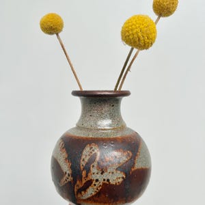 May include: A small, round ceramic vase with a dark brown and gray speckled glaze. The vase features a decorative pattern in a lighter brown color. Three yellow, spherical flowers are arranged in the vase, creating a simple, natural aesthetic.