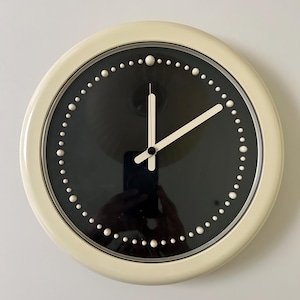 May include: Cream-colored wall clock with a black face and white hands. The clock has a circular design with white dot hour markers. The hands are white and show the time. The clock is mounted on a white wall.