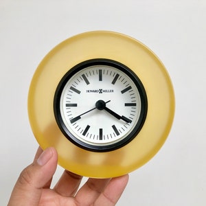 May include: A yellow circular clock with black hands and a white face. The clock face has black Roman numeral hour markers and the text "Howard Miller" printed on it.