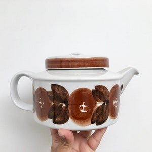 May include: A white ceramic teapot with a brown lid and a brown floral pattern. The teapot has a rounded body and a spout on one side.