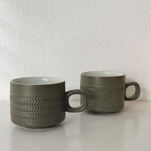May include: Two ceramic mugs with a matte olive-green exterior and a smooth white interior. Each mug features a textured band around the lower half with a repeating herringbone pattern and a small, square-shaped handle on the side.