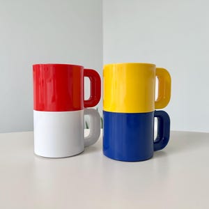 May include: Two stacked ceramic mugs. One mug is red on top and white on the bottom, with a red handle. The other mug is yellow on top and blue on the bottom, with a yellow handle. The mugs are on a white surface.