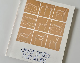 May include: A book cover featuring a grid of white line drawings of chairs on a tan background. The title "alvar aalto furniture" is in blue script. The cover is slightly angled, showing a white background.