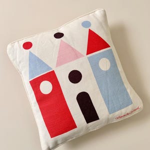 May include: A square off-white throw pillow featuring a graphic print of three stylized buildings in red, pink, and light blue with circular and arched details. The signature Alexander Girard is printed in red cursive in the bottom right corner.