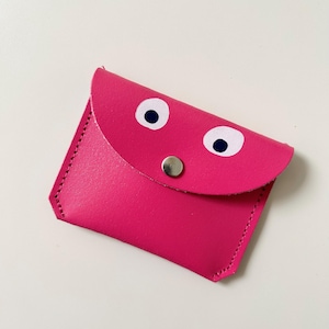 Peut inclure: Un portefeuille en cuir rose vif avec un rabat et un bouton-pression argenté. Le rabat est orné de deux cercles blancs avec des pupilles noires, ressemblant à des yeux. Le portefeuille est cousu avec du fil rose.