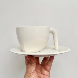 May include: A cream-colored teacup and saucer set. The cup has a unique angular design and a straight handle. The saucer is round and flat. The set is held in a hand against a plain white background.
