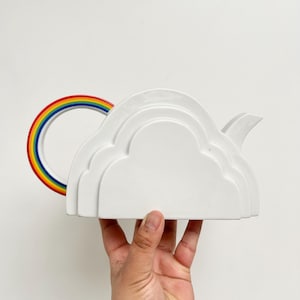 May include: A white ceramic teapot in the shape of a cloud, with a rainbow handle. The teapot has a spout and is held against a white background. The rainbow handle features red, orange, yellow, green, blue, and purple.