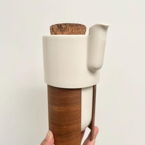 May include: A white ceramic creamer with a wooden base and a cork stopper. The creamer has a spout and a handle. The wooden base is made of dark brown wood. The creamer is held in a hand against a white background.