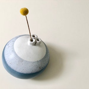 May include: A round, blue and white ceramic vase with three openings for flowers. A single yellow dried flower is in the vase.