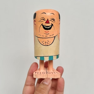 May include: A wooden figurine of a smiling man with a bald head, painted with a peach skin tone. The figure has black hair, a red nose, and a wide, toothy grin. The figure is wearing a beige top and blue and white striped shorts. The figurine is standing on two feet.