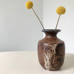 May include: A brown ceramic vase with white and brown swirling patterns. The vase has a narrow neck and a wide base. Two yellow dried flowers are in the vase.