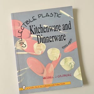 May include: A softcover book titled Collectible Plastic Kitchenware and Dinnerware 1935-1965 by Michael J. Goldberg. The light blue cover features illustrations of pink plates, cream-colored cups, and white cutlery arranged in a scattered pattern.
