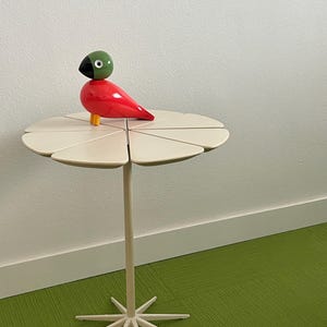 May include: A decorative side table with a cream-colored, flower-shaped top and a starburst base. A red and green bird figurine sits on the table. The table is approximately 24 inches tall.