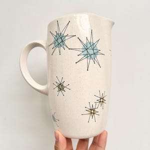 May include: A cream-colored ceramic pitcher with a speckled finish, featuring a pattern of black starburst lines overlaid on light blue and pale yellow circular shapes. The pitcher has a curved handle and a simple spout for pouring liquids.