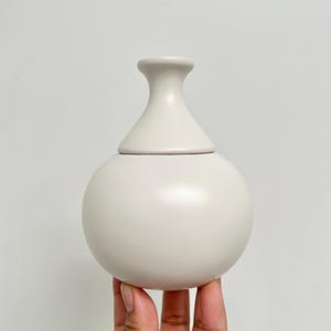 May include: A matte light beige ceramic vessel featuring a spherical body and a narrow, flared neck. The object has a smooth, uniform finish with a distinct horizontal seam where the neck meets the rounded base, creating a clean, solid silhouette.