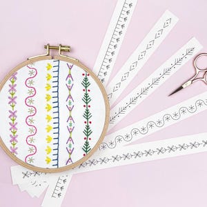 May include: A wooden embroidery hoop with colorful floral and geometric patterns stitched on white fabric. Several paper strips with black line designs and a pair of gold scissors are arranged on a pink background, perfect for craft projects.
