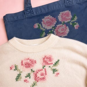 May include: A cream-colored knit sweater with a ribbed crew neckline and a cross-stitch floral pattern in pink and green, positioned in front of a navy blue canvas tote bag featuring an identical pink and green cross-stitch floral design.