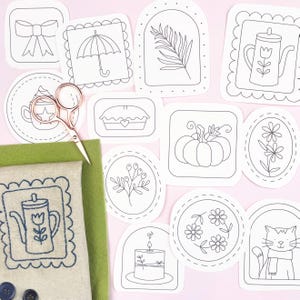 May include: A selection of embroidery templates with designs such as a teapot, pumpkin, and floral patterns. A pair of rose gold scissors and green fabric are also visible. The patterns are printed on white paper with black outlines.