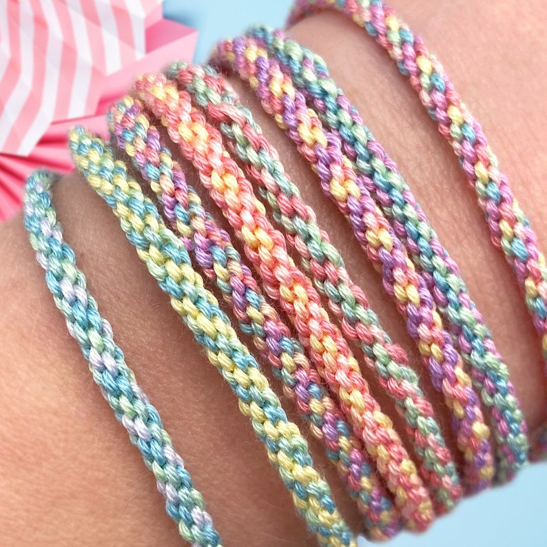 May include: A set of colorful friendship bracelets in pastel shades of blue, green, yellow, and pink. The bracelets are made with a woven pattern and are stacked on a person's wrist.