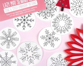 May include: A collection of white, circular stick and stitch embroidery patterns featuring black snowflake designs. The image includes a red felt Christmas tree and a pink card that reads "LAZY MAY SEWING CLUB STICK & STITCH EMBROIDERY PATTERNS".