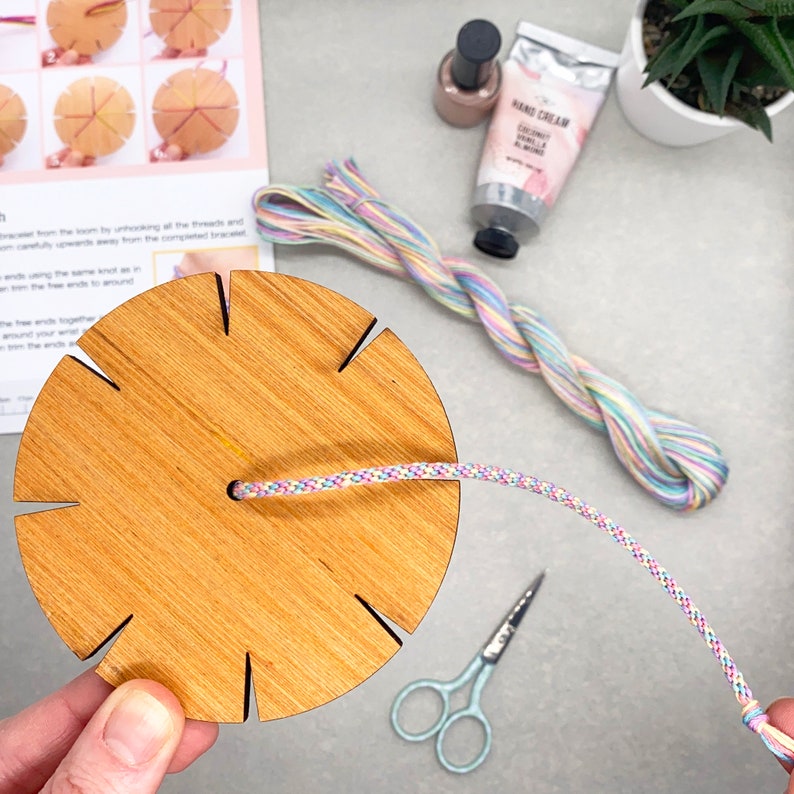 May include: A wooden bracelet making loom with a rainbow colored string threaded through it. The loom has eight slots for threading the string.