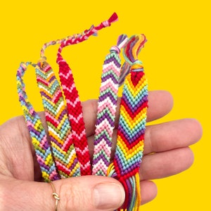 A hand holding multiple handmade friendship bracelets in bright colours. Perfect for beginners, this DIY bracelet making kit includes embroidery thread and instructions.