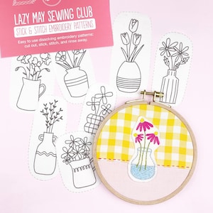 May include: A craft kit with embroidery patterns. The kit includes a wooden embroidery hoop with yellow gingham fabric, a pink fabric base, and a floral design. Several stick and stitch embroidery patterns are also included, along with a pink card that reads "LAZY MAY SEWING CLUB".