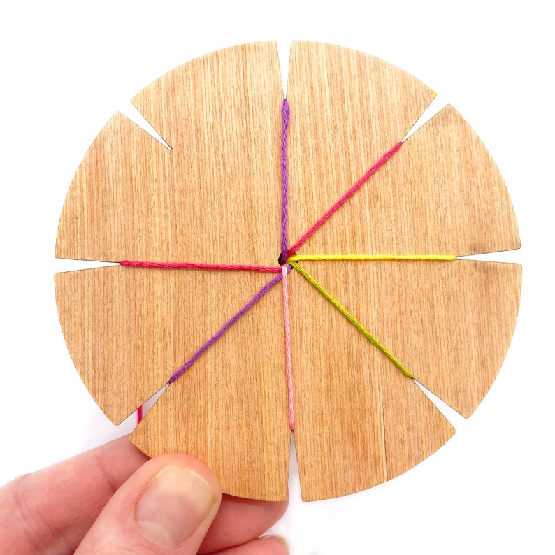 May include: A wooden loom with six slots for string. The strings are pink, purple, yellow, and red. The strings are tied together in the center of the loom.