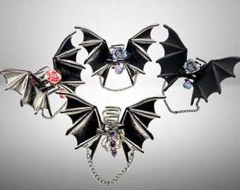 Könnte beinhalten: Fünf Fledermausflügelringe in Silber und Schwarz. Jeder Ring hat detaillierte Fledermausflügel, ein dekoratives Zentrum und eine Kette. Einige haben Rosen- oder Edelsteinakzente. Ein Gothic-inspiriertes Accessoire.