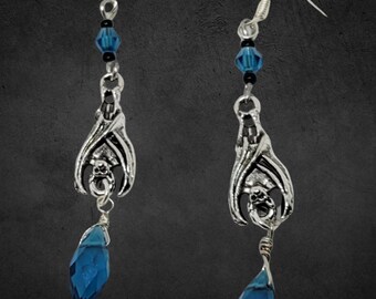 Puede incluir: Un par de pendientes de plata con diseño de murciélago y una gota de cristal azul oscuro. Los pendientes tienen un cierre de gancho de plata.