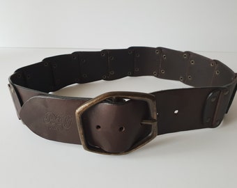 May include: A wide, dark brown leather belt with a large, rectangular brass buckle. The belt features a series of rectangular segments connected by rivets. The D&G logo is embossed on the leather.