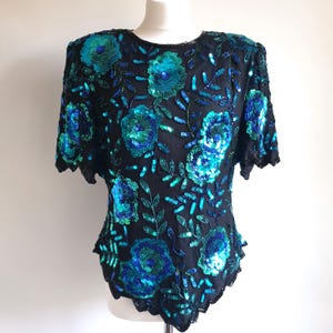 Puede incluir: Top negro de manga corta con adornos florales de lentejuelas en tonos turquesa y azul. Las lentejuelas forman grandes patrones de flores y diseños de hojas en la prenda. El top se muestra en un maniquí.