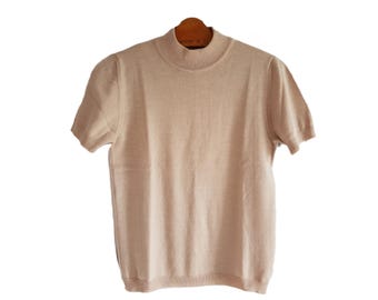 May include: A short-sleeved, light tan knit top with a mock turtleneck. The shirt is hanging on a wooden hanger against a white background. The knit fabric has a subtle texture.